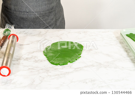 A close, eye-level view capturing hands using a rolling pin to flatten green dough on a marble surface. This step prepares the dough for cutting out shapes for the Green Gingerbread Cookies recipe. A close, eye-level view capturing hands using a rolling pin to flatten green dough on a marble surface. This step prepares the dough for cutting out shapes for the Green Gingerbread Cookies recipe. 130544466