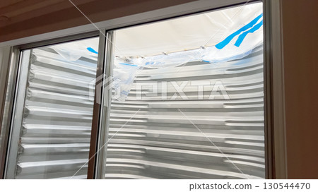 A metal basement window well is viewed from the inside through a sliding glass window. The window is partially covered with white plastic for protection during construction or renovation. 130544470