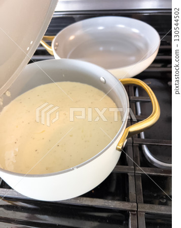 A light-colored creamy sauce is being stirred in a white pot with golden handles. The pot sits on a stove, and the texture of the sauce appears smooth and velvety. The kitchen lighting is bright and A light-colored creamy sauce is being stirred in a white pot with golden handles. The pot sits on a stove, and the texture of the sauce appears smooth and velvety. The kitchen lighting is bright and 130544502