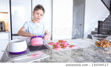 With focused attention, this budding culinary artist dips pretzels into a pot of melted chocolate, creating sweet delights in the warmth of a well-lit home kitchen. 130544517