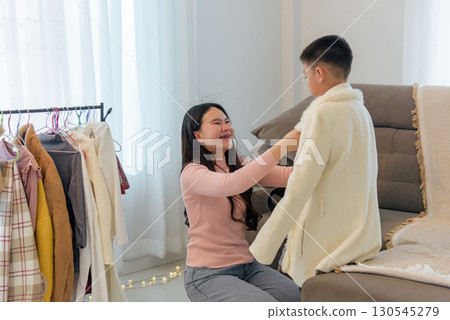 Woman helps a child to put on a warm winter coat, a clothing rack shows more winter attire, as they prepare for the cold day ahead indoors. 130545279