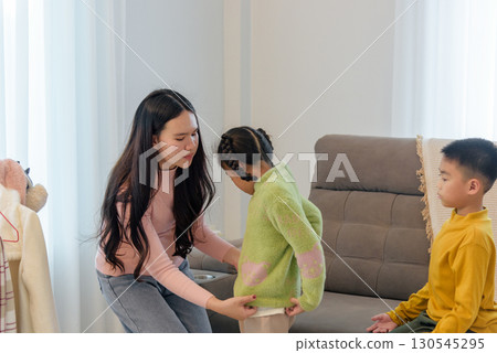 During a winter day an Asian woman is helping a child with her green sweater, another child sits on a couch in a yellow sweater, a warm scene for family clothing needs. 130545295