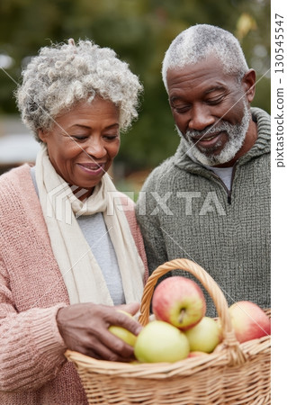 Happy senior couple harvesting apples into a wicker basket on a crisp autumn day, enjoying time together in the orchard Happy senior couple harvesting apples into a wicker basket on a crisp autumn day, enjoying time together in the orchard 130545547