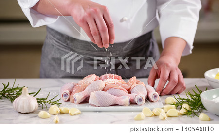 Chef seasoning raw chicken drumsticks with salt, preparing meat with garlic and rosemary on kitchen counter. Cooking preparation scene. 130546010