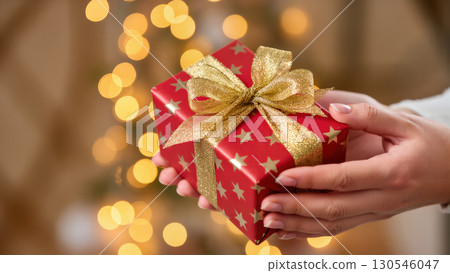 Christmas gift in hands with red wrapping paper, gold stars and glitter ribbon, festive bokeh lights in background Christmas gift in hands with red wrapping paper, gold stars and glitter ribbon, festive bokeh lights in background 130546047
