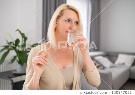 Middle aged woman holding pill between fingers and glass of water, focused expression, sitting in cozy living room  130547031
