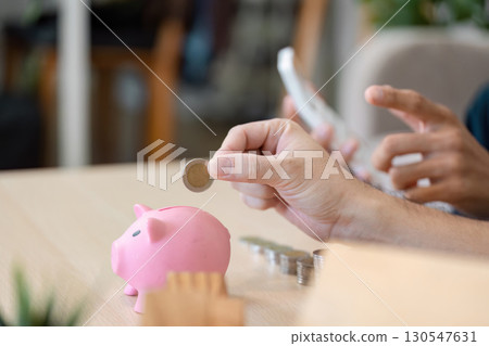 LGBTQ couple adding a coin to a pink piggy bank while using a smartphone for budgeting in their cozy living room. Emphasis on financial planning and teamwork. 130547631