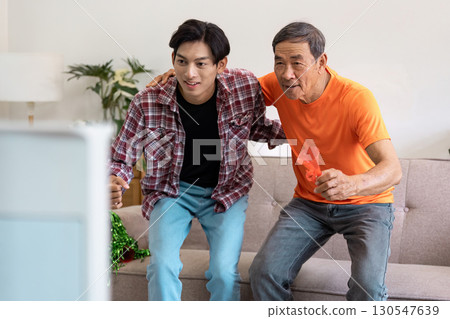 Excited Asian father and son cheering together while watching a soccer game at home, showcasing family bonding and enthusiasm. Excited Asian father and son cheering together while watching a soccer game at home, showcasing family bonding and enthusiasm. 130547639