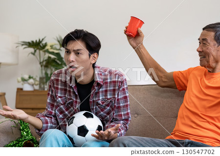 Surprised Asian son reacting to a soccer play while his father cheers at home, showcasing family emotions and sports dynamics. 130547702
