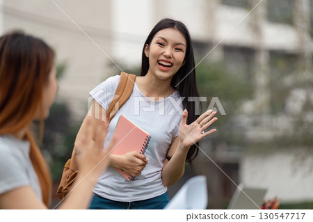 Diverse university student smiling and waving while holding a notebook outdoors. 130547717