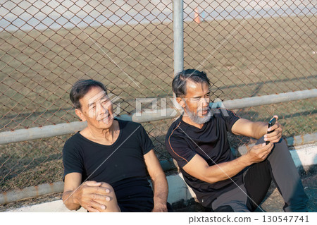 Content elderly men relaxing together and using a smartphone after a workout on the track. Emphasizing friendship, wellness, and active aging. Content elderly men relaxing together and using a smartphone after a workout on the track. Emphasizing friendship, wellness, and active aging. 130547741