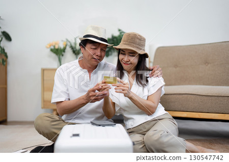 Affectionate senior couple planning their travel while looking at a credit card and sitting beside a suitcase at home. Affectionate senior couple planning their travel while looking at a credit card and sitting beside a suitcase at home. 130547742