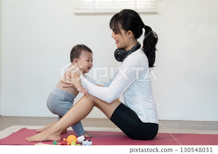 Mother encouraging her baby to stand while she sits on a yoga mat, fostering playfulness and support during a home workout session. 130548031