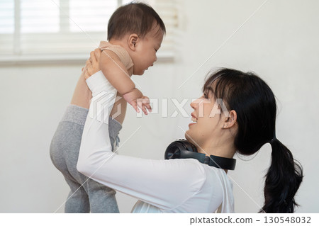 Mother lifting her baby joyfully in the air, both sharing smiles and laughter during a playful moment at home, emphasizing family love. Mother lifting her baby joyfully in the air, both sharing smiles and laughter during a playful moment at home, emphasizing family love. 130548032