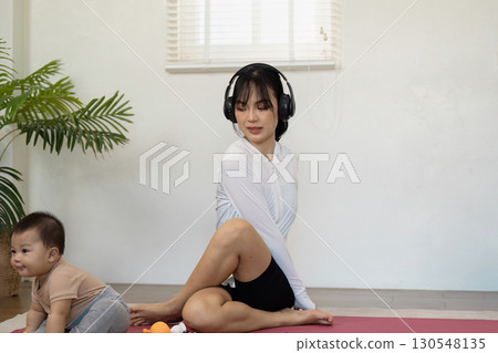 Mother practicing yoga at home with headphones while baby explores. Mother practicing yoga at home with headphones while baby explores. 130548135