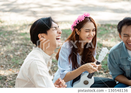 Diverse group of friends laughing and playing ukulele in a sunny park. 130548136