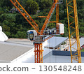 Large orange construction crane with glass cabin operating beside metal-roof buildings, set against lush green trees, symbolizing urban development, engineering progress, and industrial growth 130548248