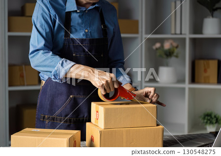 E-commerce worker packing boxes for online sales with tape in a workspace. 130548275