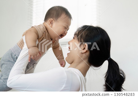 Mother joyfully lifting her baby in the air, sharing laughter and love during a playful moment at home, emphasizing family connection. Mother joyfully lifting her baby in the air, sharing laughter and love during a playful moment at home, emphasizing family connection. 130548283