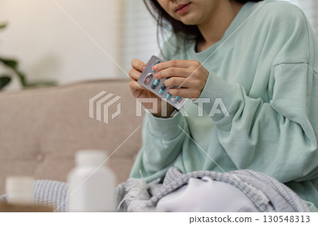 Young woman holding a blister pack of medication while resting on a couch, emphasizing health management and self-care. 130548313