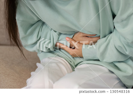 Young woman experiencing abdominal discomfort while sitting on a couch, highlighting health issues and self-awareness. Young woman experiencing abdominal discomfort while sitting on a couch, highlighting health issues and self-awareness. 130548316