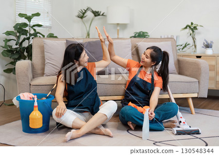 Two women giving high-fives while seated on a couch, surrounded by cleaning supplies and a bucket. 130548344
