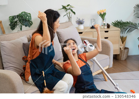 Two women celebrating their cleaning achievements while sitting on a couch, one with a vacuum cleaner and the other with a feather duster. 130548346