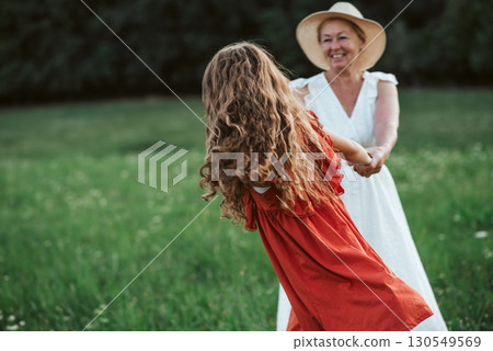 Grandma and girl holding hands and dancing in the middle of meadow. Grandma and girl holding hands and dancing in the middle of meadow. 130549569