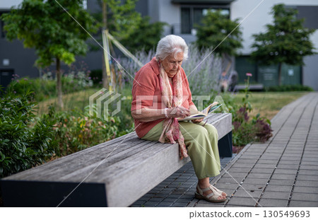 Elderly woman reading a book outdoors in urban garden. Elderly woman reading a book outdoors in urban garden. 130549693