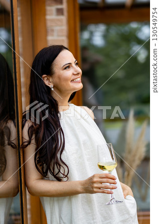 Smiling woman with glass of white wine, standing on terrace. 130549754