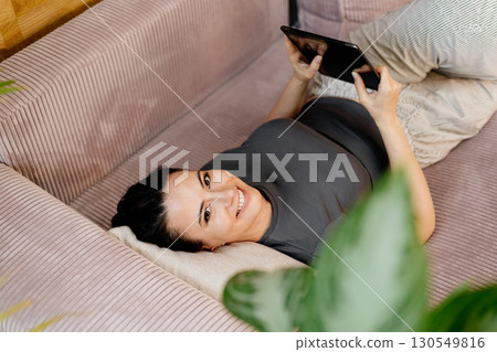 Relaxed woman taking a break, lying on sofa and scrolling on tablet. 130549816