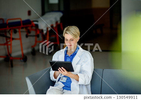 Young female doctor with short hair in sitting on bench in hospital corridor, holding tablet. 130549911