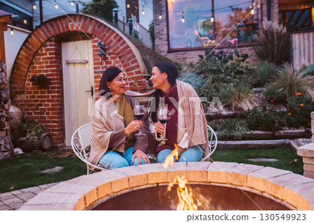 Elderly mother and adult daughter having glass of wine together, sitting by fire pit. Elderly mother and adult daughter having glass of wine together, sitting by fire pit. 130549923