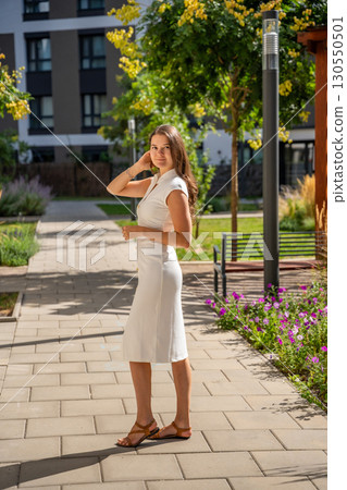Young woman wearing a white midi dress walking outdoors in Old Money style. Concept of timeless fashion, graceful femininity and understated elegance in modern lifestyle. 130550501