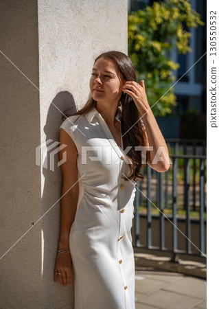 Young woman wearing a white midi dress walking outdoors in Old Money style. Concept of timeless fashion, graceful femininity and understated elegance in modern lifestyle. Young woman wearing a white midi dress walking outdoors in Old Money style. Concept of timeless fashion, graceful femininity and understated elegance in modern lifestyle. 130550532