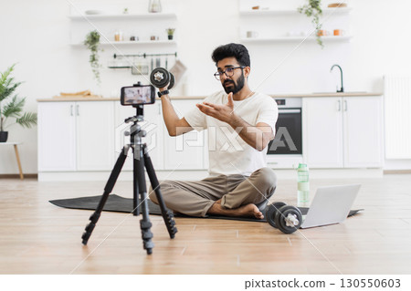 Man sitting on yoga mat recording fitness blog at home using dumbbell and tripod camera setup. Concept highlights health, fitness, and modern lifestyle promoting home-recorded fitness video 130550603