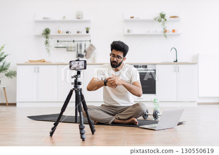 Caucasian adult man in casual outfit recording sports-related vlog discussing smart fitness devices. Room setting is a modern kitchen with exercise equipment visible. 130550619