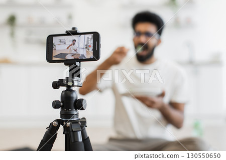 Young adult man sits in kitchen recording video about healthy eating habits. Scene shows him eating and using smartphone camera for vlog, promoting balanced diet and modern lifestyle. Young adult man sits in kitchen recording video about healthy eating habits. Scene shows him eating and using smartphone camera for vlog, promoting balanced diet and modern lifestyle. 130550650