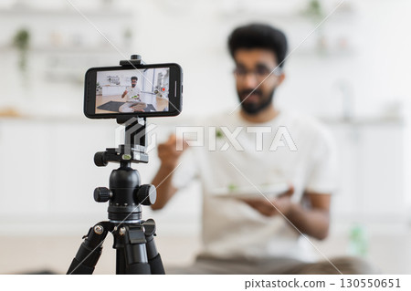 Young adult man sits in kitchen recording video about healthy eating habits. Scene shows him eating and using smartphone camera for vlog, promoting balanced diet and modern lifestyle. Young adult man sits in kitchen recording video about healthy eating habits. Scene shows him eating and using smartphone camera for vlog, promoting balanced diet and modern lifestyle. 130550651