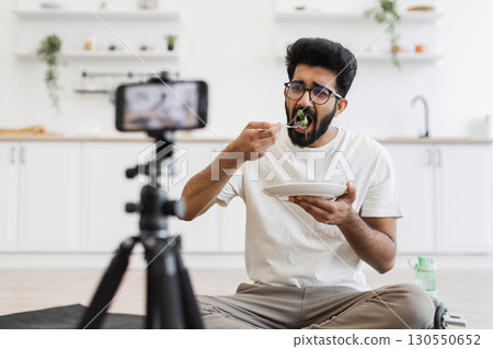 Young adult man sits in kitchen recording video about healthy eating habits. Scene shows him eating and using smartphone camera for vlog, promoting balanced diet and modern lifestyle. 130550652