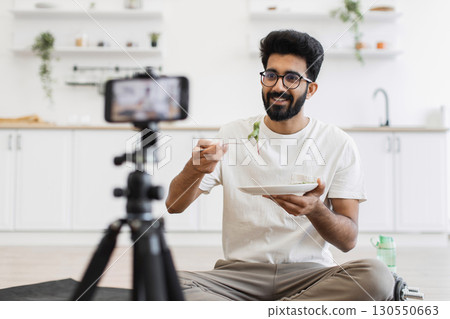 Young adult man sits in kitchen recording video about healthy eating habits. Scene shows him eating and using smartphone camera for vlog, promoting balanced diet and modern lifestyle. Young adult man sits in kitchen recording video about healthy eating habits. Scene shows him eating and using smartphone camera for vlog, promoting balanced diet and modern lifestyle. 130550663