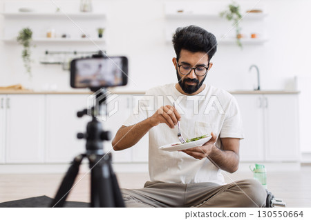 Middle age man sitting on yoga mat in bright kitchen recording healthy eating blog with smartphone focused on healthy lifestyle wellness capturing audience interest. Laptop food props enhance setup. Middle age man sitting on yoga mat in bright kitchen recording healthy eating blog with smartphone focused on healthy lifestyle wellness capturing audience interest. Laptop food props enhance setup. 130550664