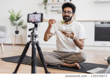 Man sitting cross-legged in kitchen filming a video about healthy eating for viewers. Includes indoor background, white shirt, home setting, casual atmosphere. Man sitting cross-legged in kitchen filming a video about healthy eating for viewers. Includes indoor background, white shirt, home setting, casual atmosphere. 130550671