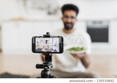 Young adult man sitting indoors recording vlog discussing healthy eating, focused on smartphone camera. 130550682