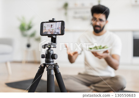 Young adult man sitting indoors recording vlog discussing healthy eating, focused on smartphone camera. Young adult man sitting indoors recording vlog discussing healthy eating, focused on smartphone camera. 130550683