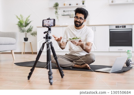 Young adult man sitting on mat in kitchen recording video about healthy eating lifestyle showing food plate to camera. 130550684