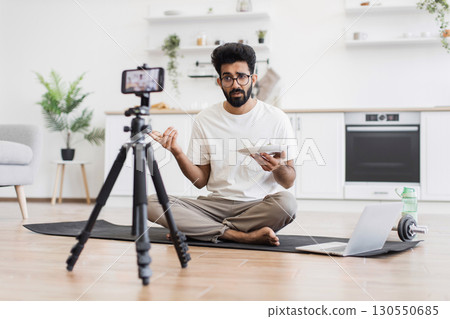 Young adult man sitting on mat in kitchen recording video about healthy eating lifestyle showing food plate to camera. 130550685