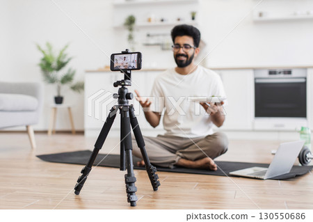 Young bearded man sitting on floor in modern apartment recording video about healthy eating. 130550686