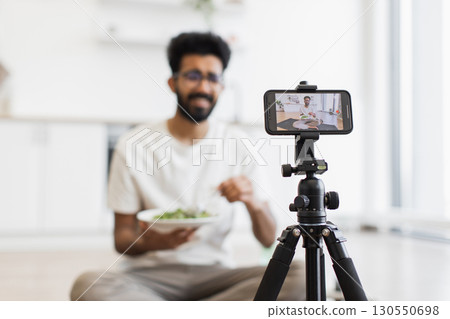 Man in his twenties wearing glasses holding bowl with salad filming video blog in bright kitchen. Video showcasing healthy eating habits, vlog recording setup with a tripod-mounted phone camera. 130550698