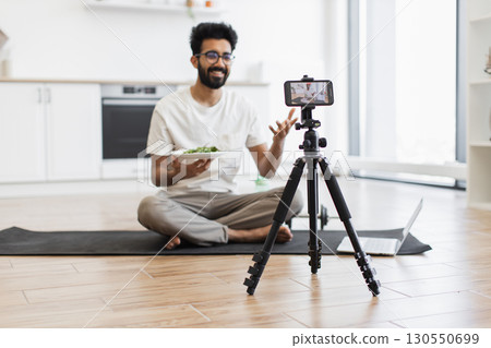Young adult man sitting cross-legged on yoga mat in modern kitchen recording video blog with smartphone and tripod holding healthy salad plate. Scene suggests health, nutrition, video blogging. Young adult man sitting cross-legged on yoga mat in modern kitchen recording video blog with smartphone and tripod holding healthy salad plate. Scene suggests health, nutrition, video blogging. 130550699